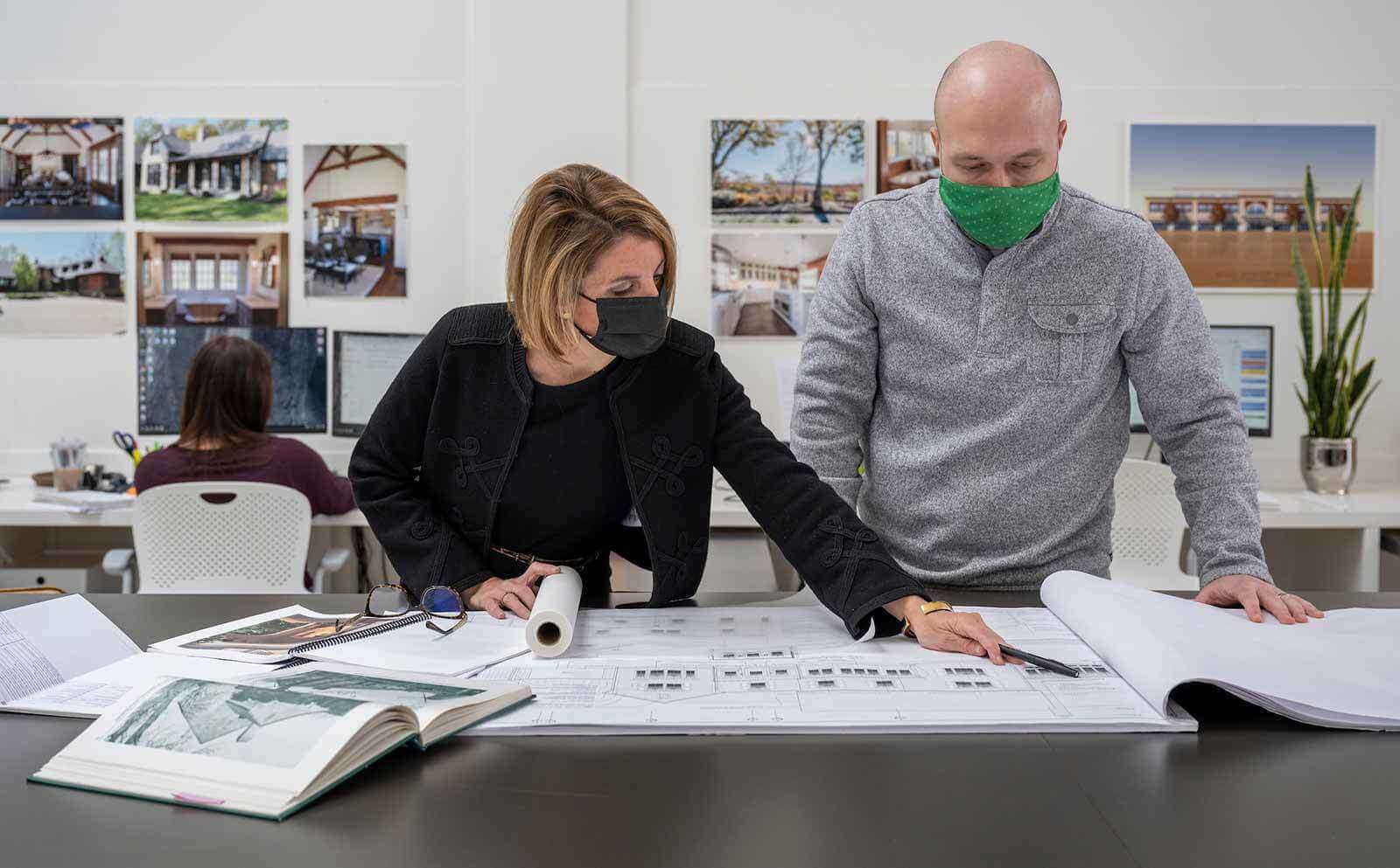 Two masked people stand at a desk looking at architecture sketches.