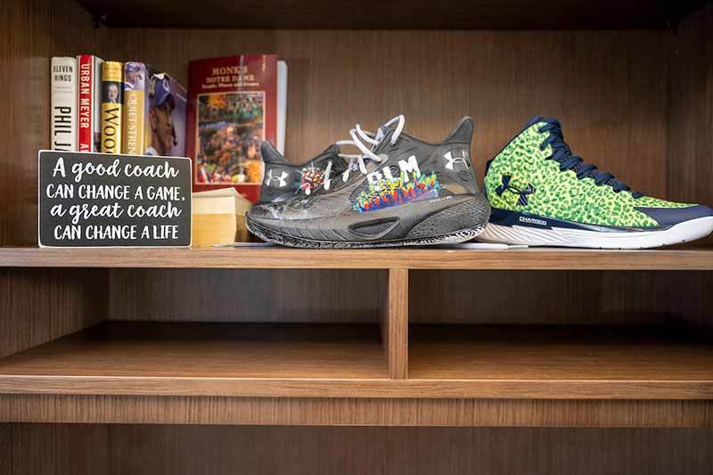 Coach Ivey's office shelf displaying books, decorated 'BLM' basketball shoes, and a wooden sign that says 'A good coach can change a game, a great coach can change a life'.